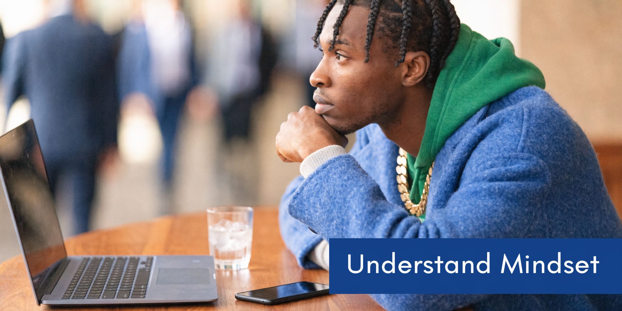 A young man with braided hair looking thoughtfully at his laptop screen while sitting at a table.