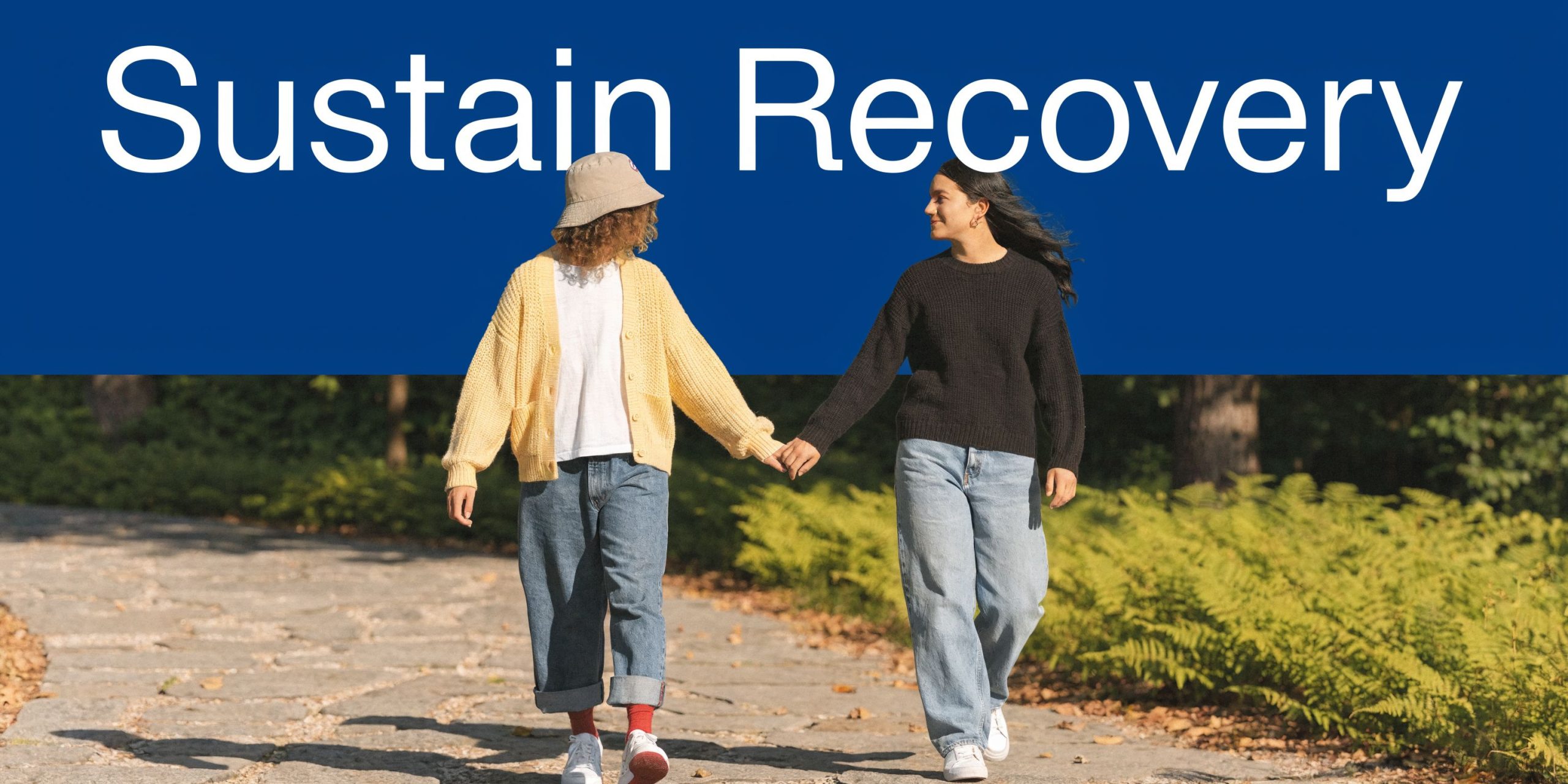 Two friends holding hands while walking on a path in a park, symbolizing recovery and emotional support.