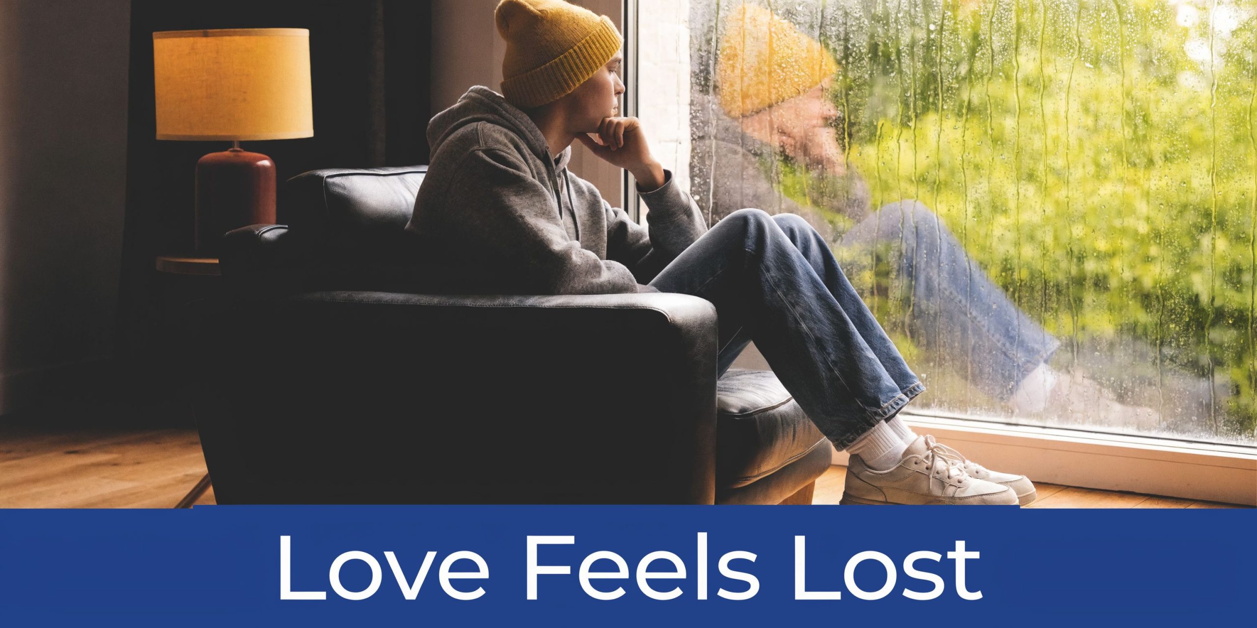 A lonely person wearing a beanie sits in a chair looking out a rainy window, feeling sad.