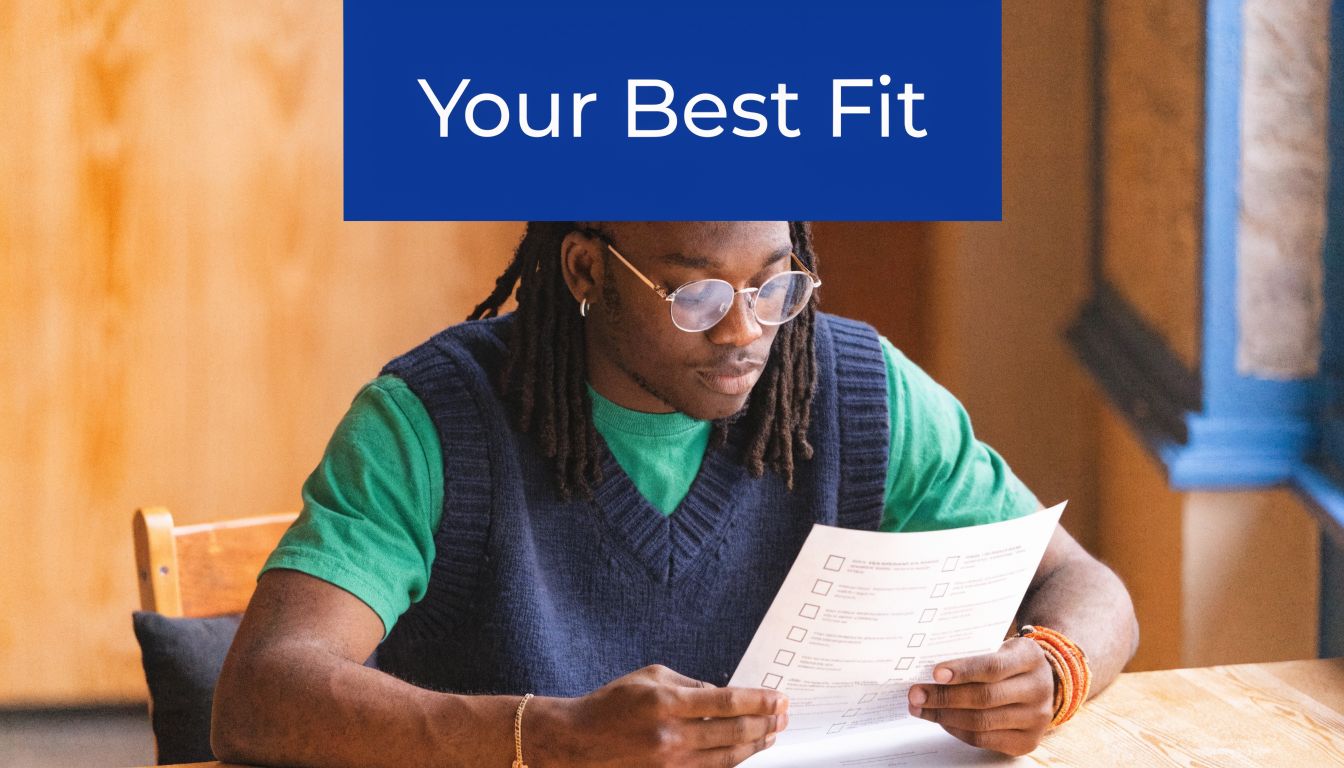 A young man with dreadlocks wearing glasses and a sweater vest reviews a document at a table.
