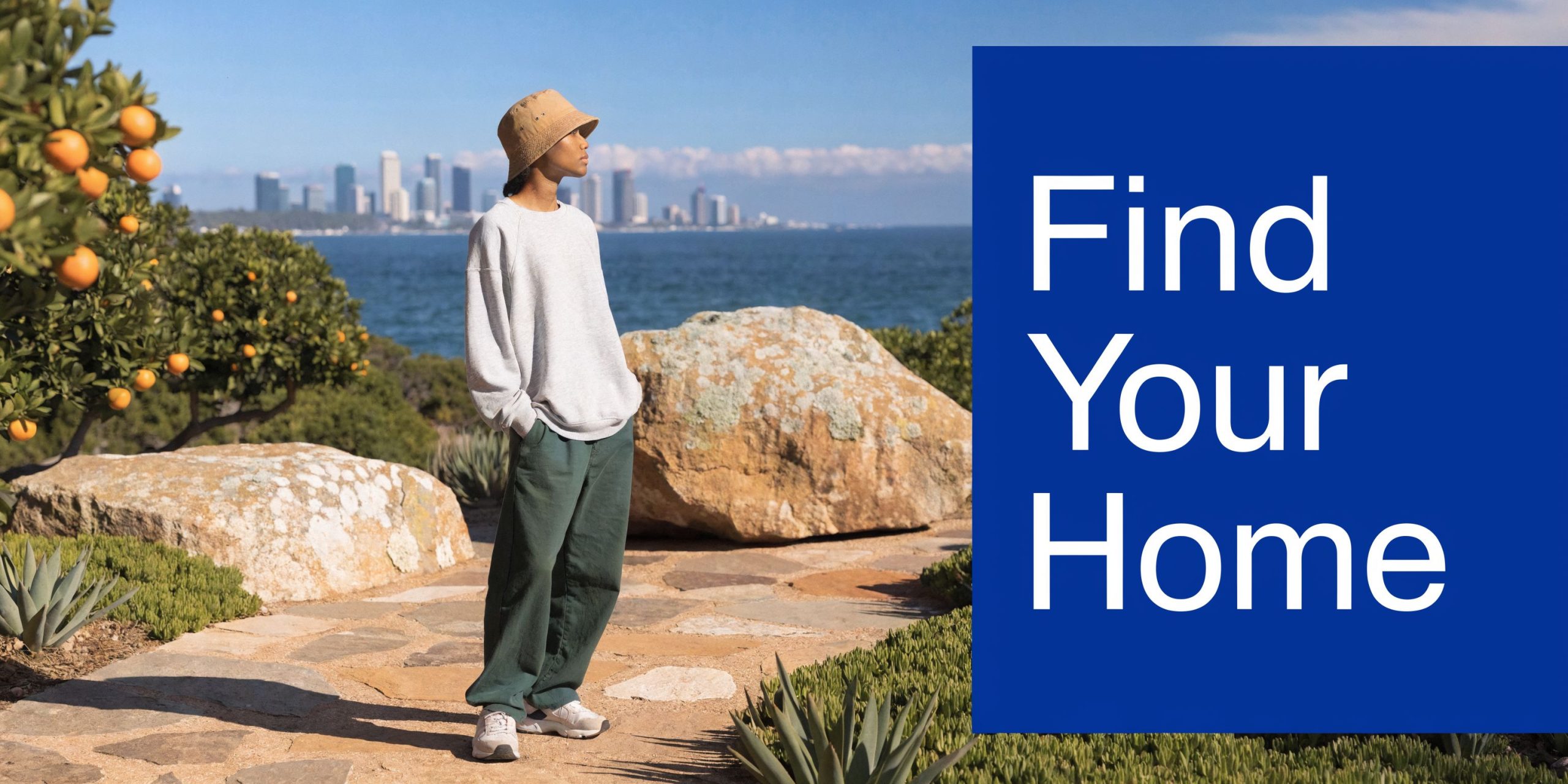 A person wearing a hat and casual clothing standing by a scenic coastal path overlooking a city.